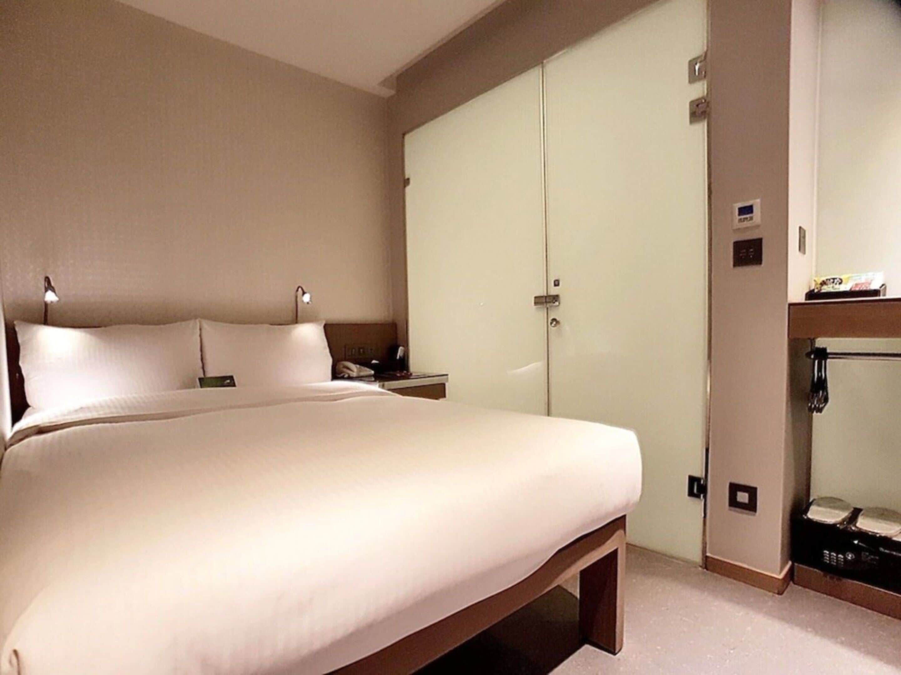 A hotel room with a large bed, a bedside table with a lamp, a wardrobe, and a small shelf with items.