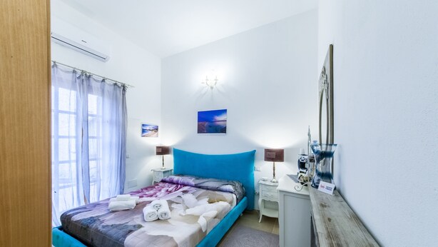 A bedroom with a bed, two bedside tables, a nightstand, a mirror, and a window with curtains. - Acuario Guest House (Oristano)