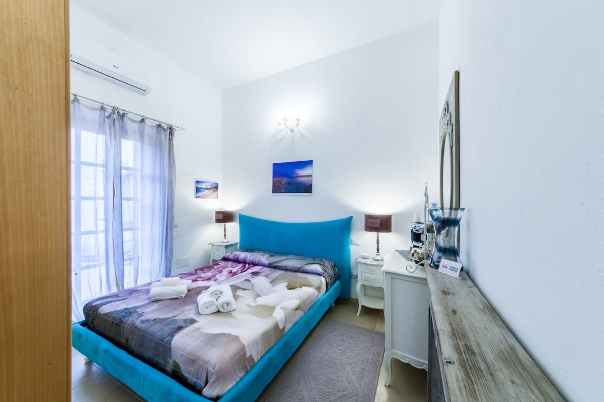 Quarto com cama, duas mesinhas de cabeceira, um criado-mudo, espelho e janela com cortinas.