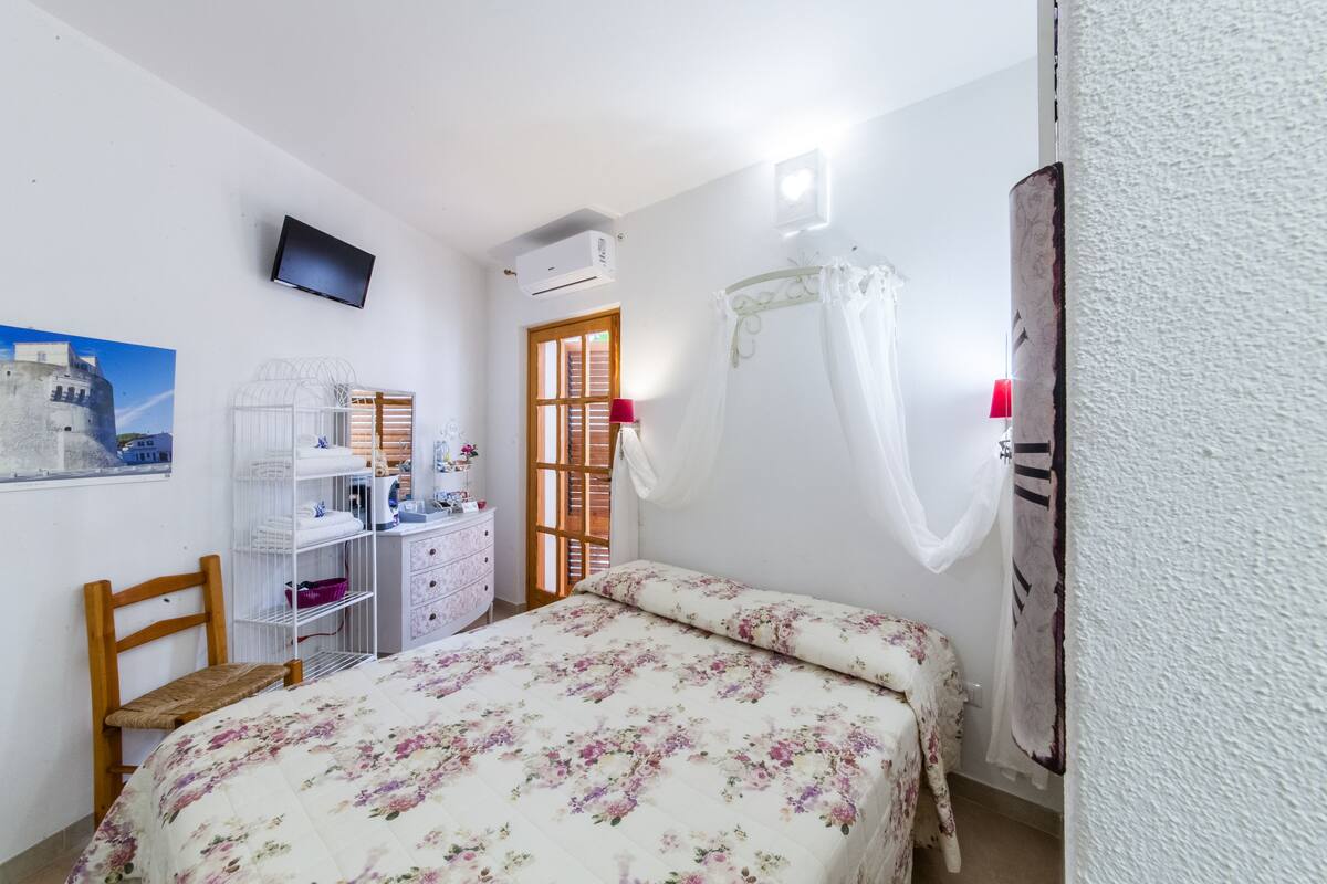 Quarto com cama, uma cadeira, escrivaninha com cadeira, televisão, ar condicionado e janela com cortinas.