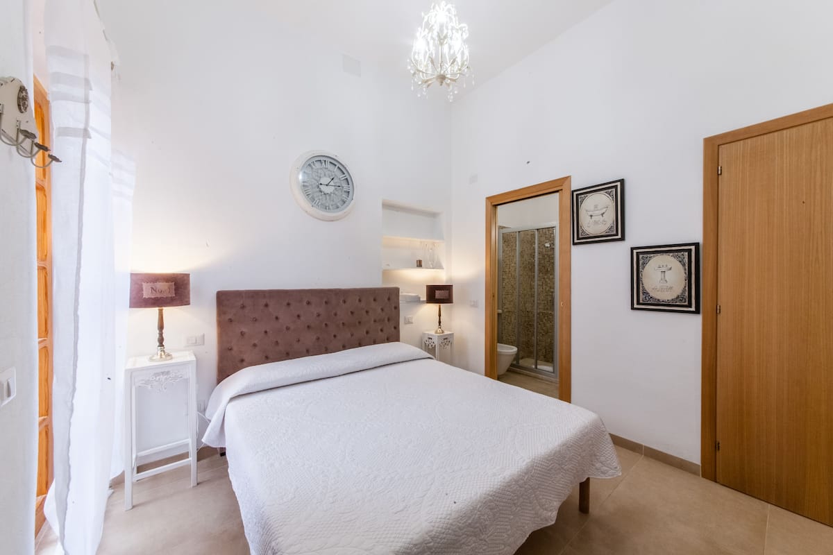Quarto com cama, relógio, espelho e porta.