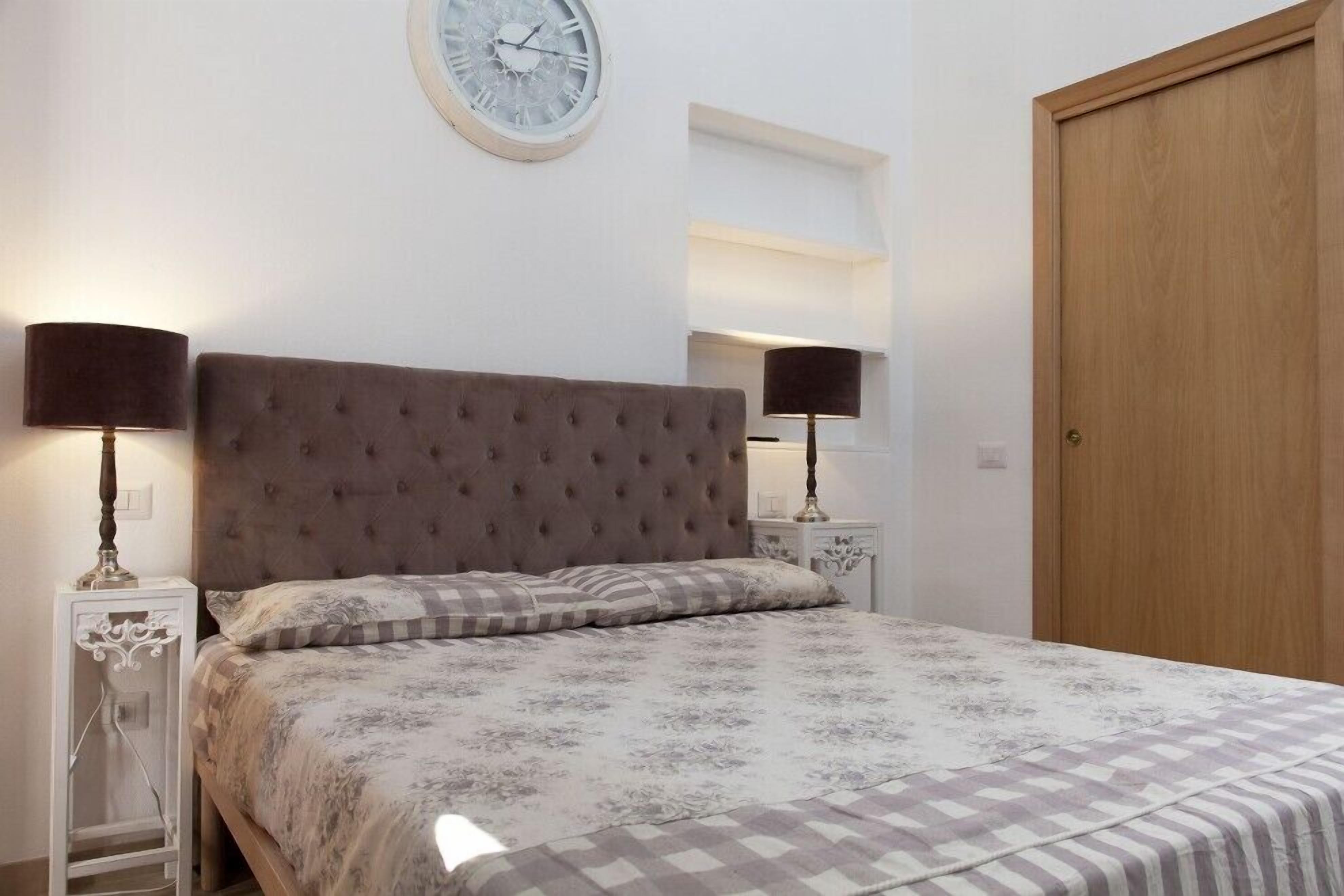 Ein Schlafzimmer mit einer gepolsterten Kopfteil, einem Bett mit gemusterten Betttextilien, zwei Nachttischen mit Lampen, einer runden Wanduhr und einer Holztür.