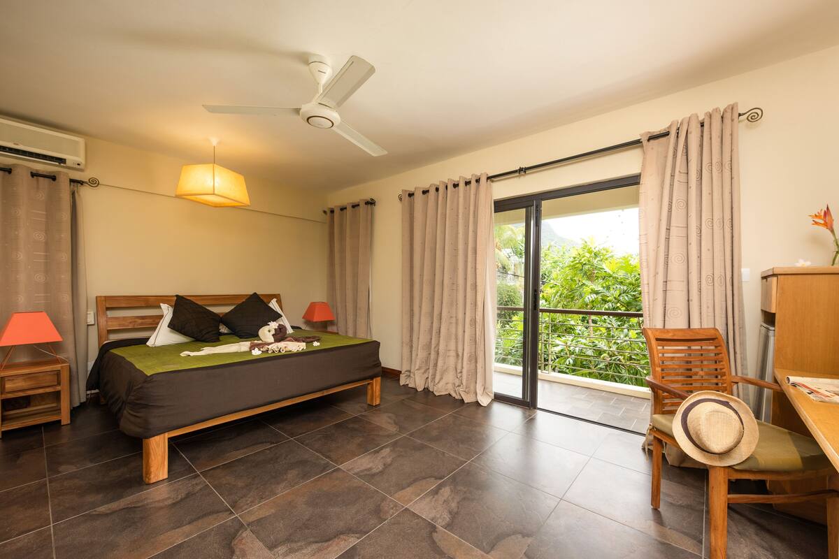 Ein Schlafzimmer mit Bett, Schreibtisch, Stuhl, Deckenventilator und Blick auf Grün durch das Fenster.