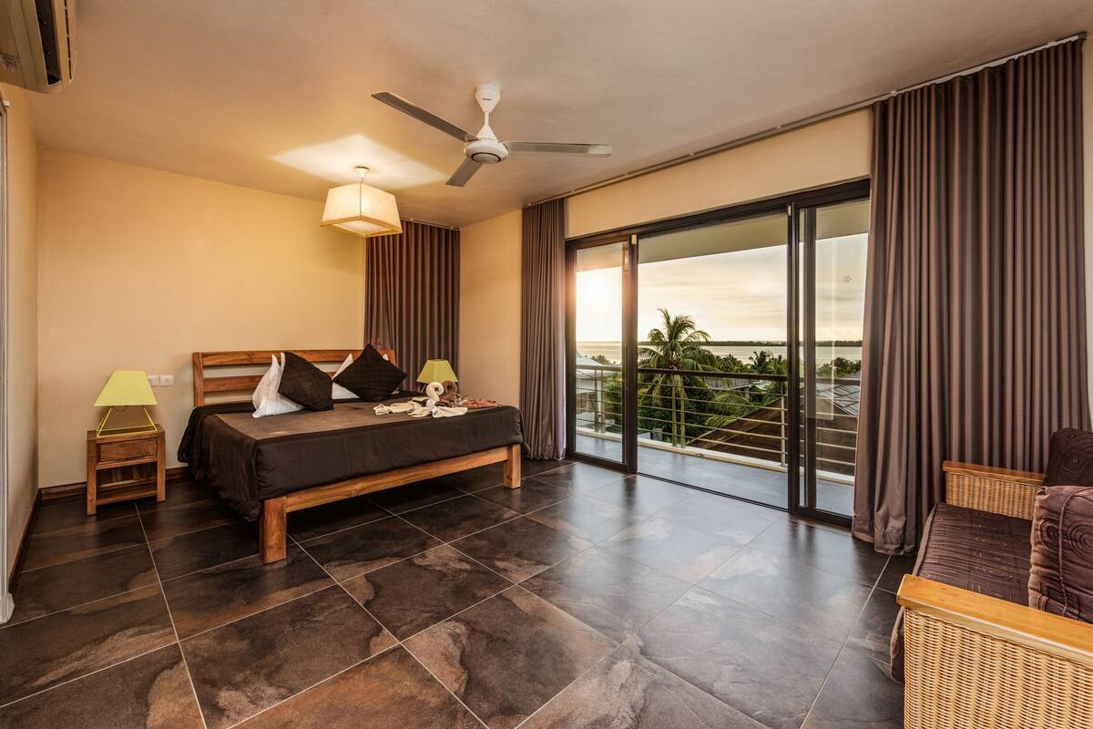 Ein Schlafzimmer mit Bett, Nachttisch, Lampe, Deckenventilator und Blick ins Freie.