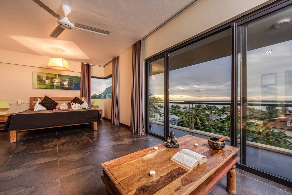 Ein modernes Hotelzimmer mit einem großen Bett, einem Holztisch mit einem Buch und einer Schale, und Blick auf die Stadt durch große Fenster.