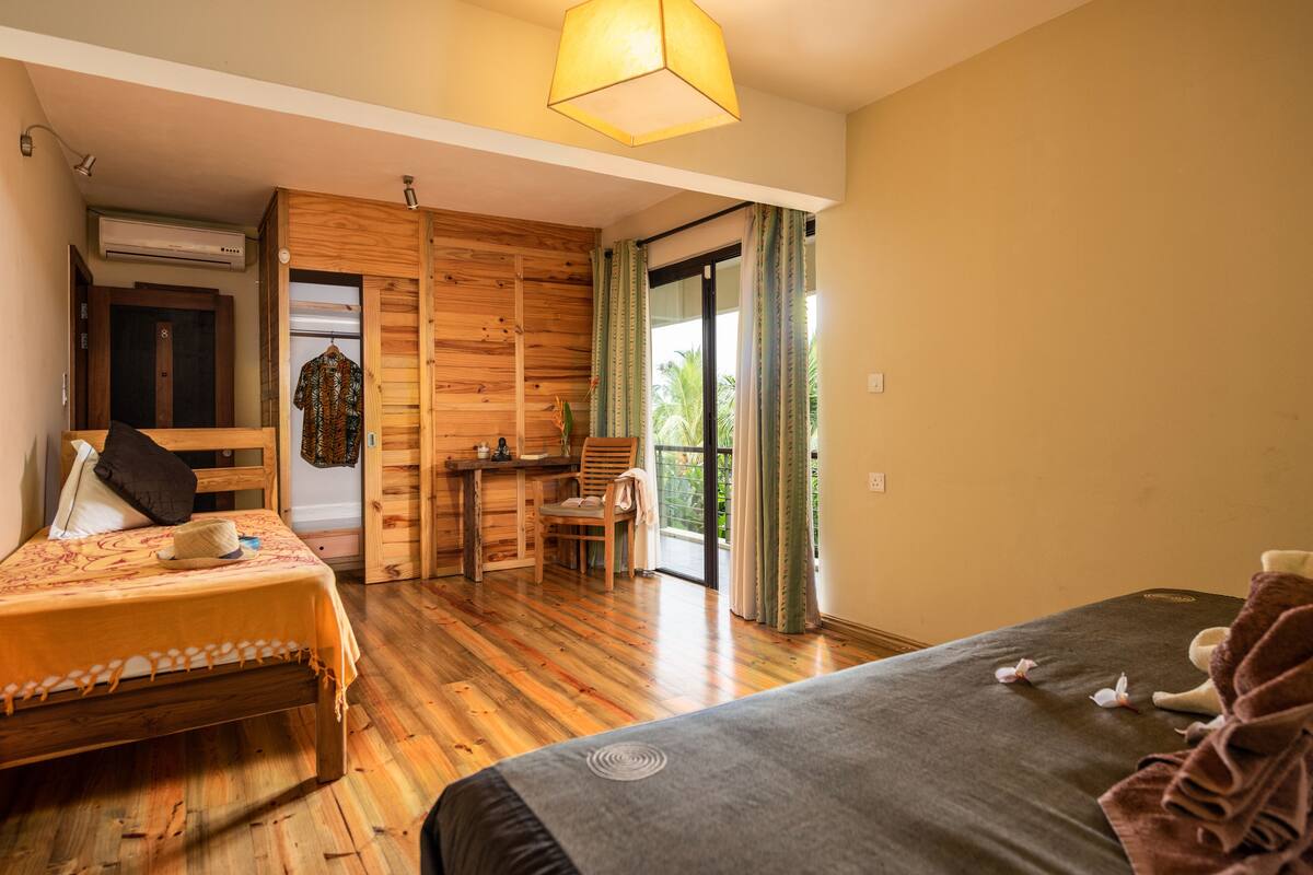 Ein Zimmer mit zwei Betten, einem Holztisch, einem Stuhl, einem Kleiderhaken und einer Glastür, die auf einen Balkon führt.