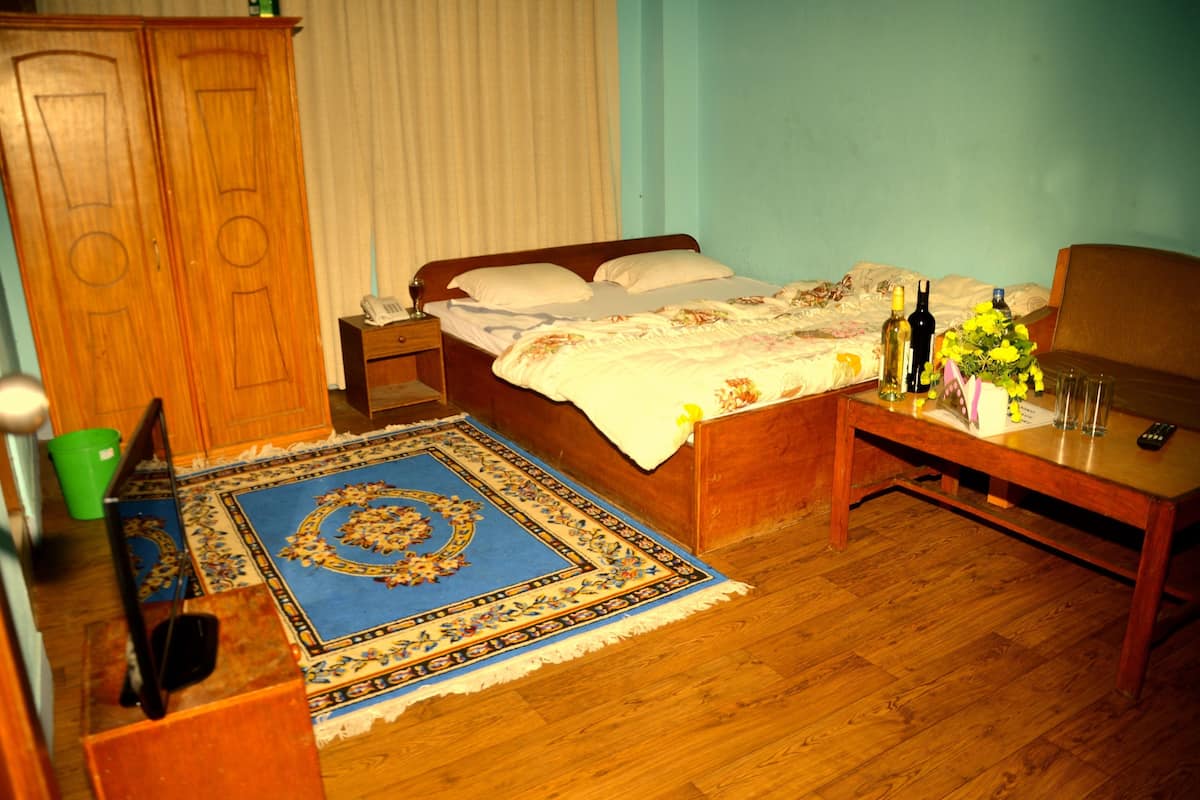 A bedroom with a wooden bed, a nightstand, a green bucket, a blue patterned rug, and a wooden table with a vase of flowers and bottles.