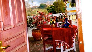 Una mesa roja con una silla, un narguile y una planta en maceta en un balcón con vistas a un pueblo y montañas.
