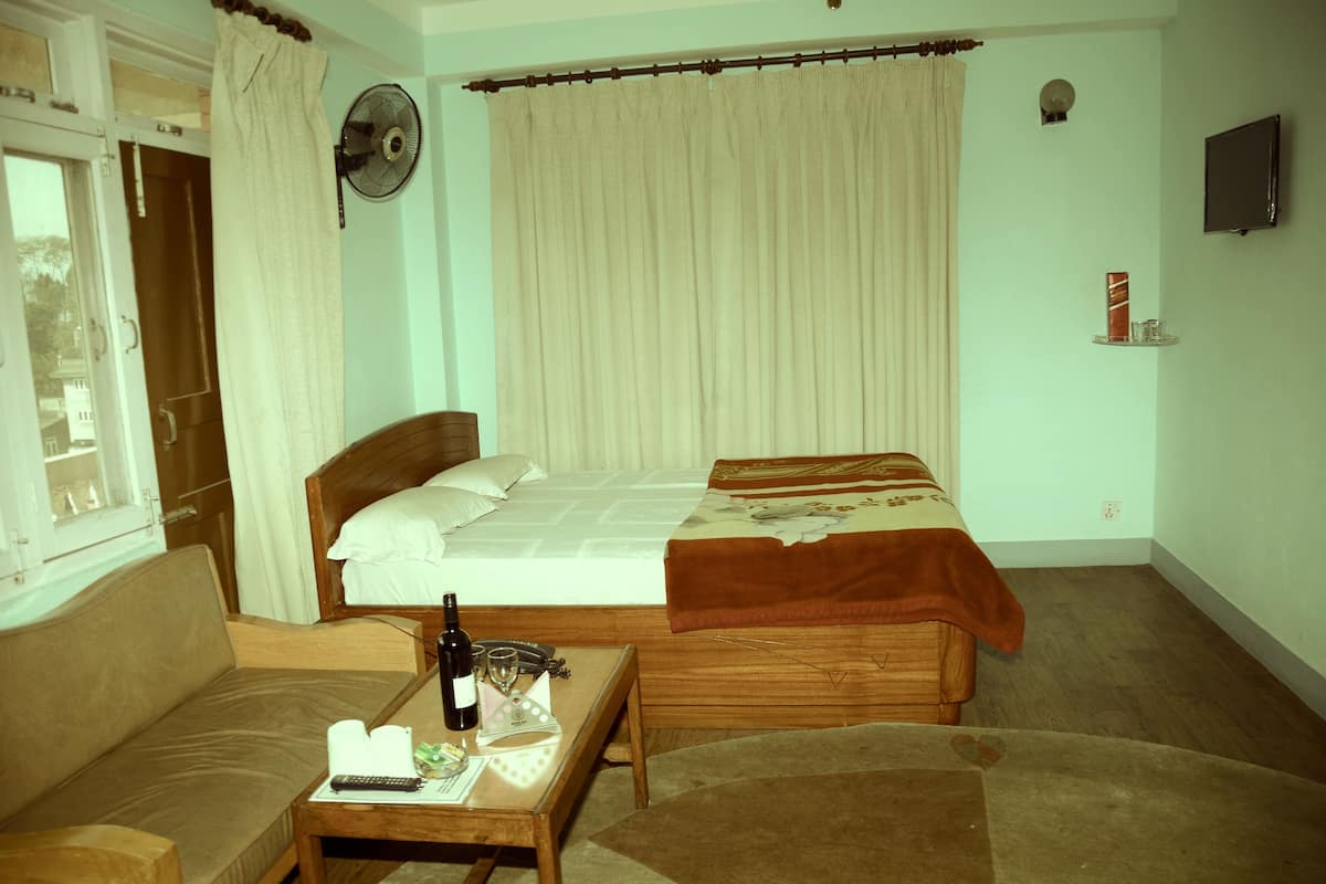 A bedroom with a bed, a wooden nightstand, a table with a bottle and glasses, a fan, and a window with curtains.