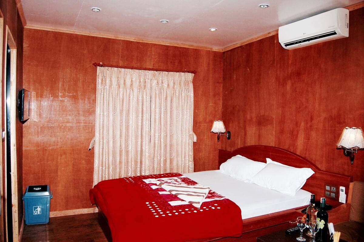 A hotel room with a wooden wall, a bed with white bedding, a red throw blanket, a nightstand with a lamp, and a trash bin.