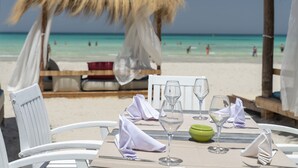A beachside dining table with white chairs, a thatched umbrella, and a view of the ocean. - Yadis Djerba Thalasso & Golf (Djerba Midun)