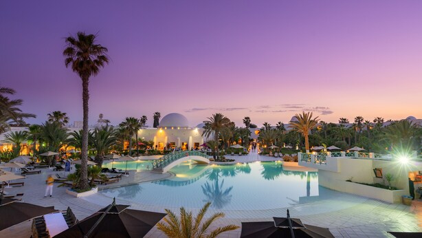 A resort pool area with palm trees, lounge chairs, umbrellas, and a dome-shaped building in the background. - Yadis Djerba Thalasso & Golf (Djerba Midun)
