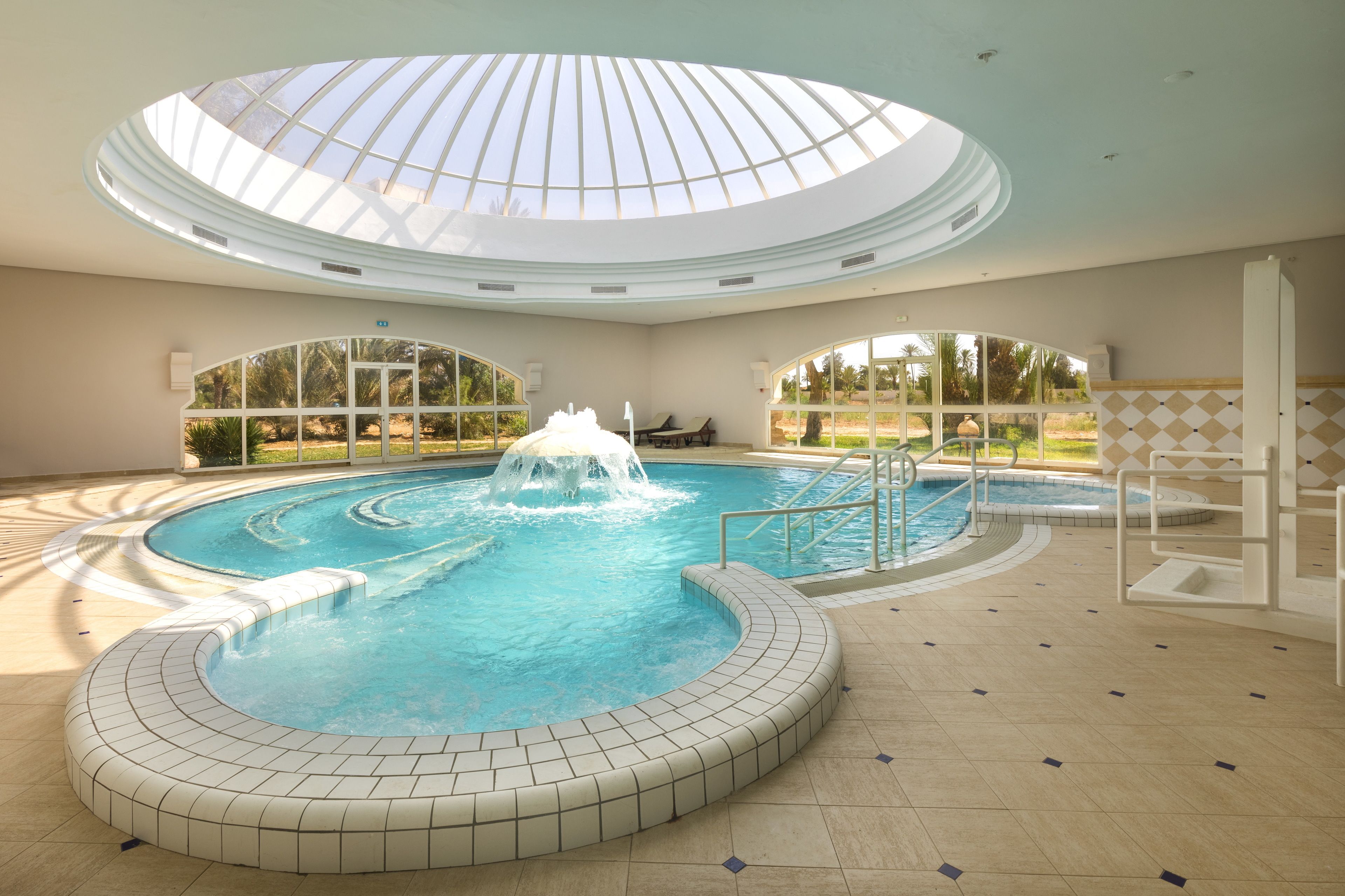 An indoor swimming pool with a central fountain, surrounded by a tiled deck and a glass ceiling.