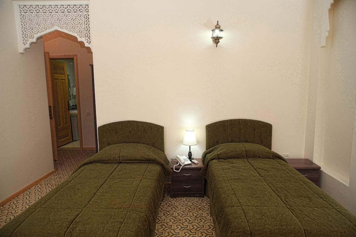 A room with two single beds, a nightstand with a lamp and phone, patterned carpet, and a decorative wall hanging.