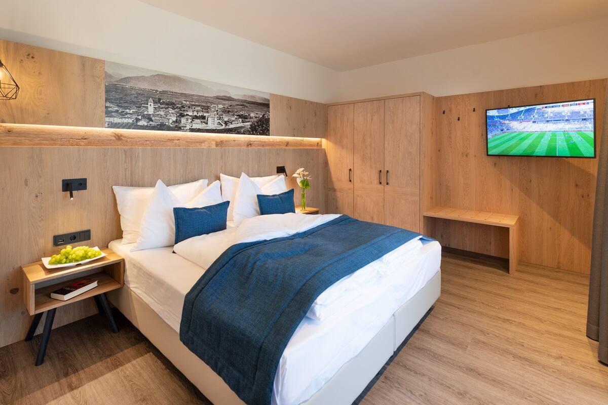 Une chambre à coucher moderne avec un lit, une tête de lit en bois, une table de chevet sur laquelle se trouve une assiette de fruits, un téléviseur fixé au mur et un tableau au-dessus du lit.