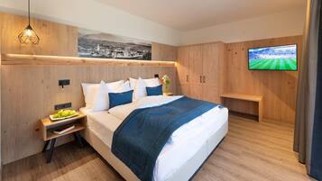 A modern bedroom with a bed, wooden headboard, a nightstand with a plate of fruit, a TV on the wall, and a painting above the bed.