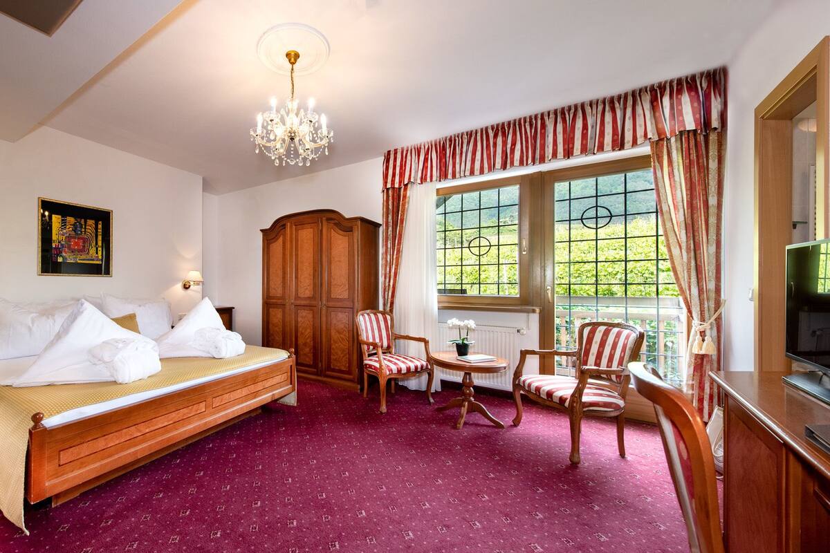 Une chambre d’hôtel équipée d’un lit, d’une télévision, d’un bureau, d’une chaise, d’une fenêtre avec des rideaux et d’un lustre.