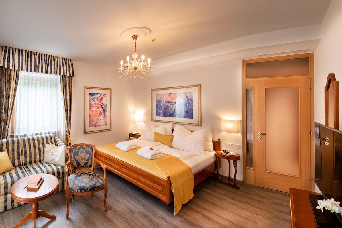 Une chambre d’hôtel avec un lit, un canapé, une chaise, une petite table et un bureau. Il y a deux tableaux au mur, un lustre et une porte.