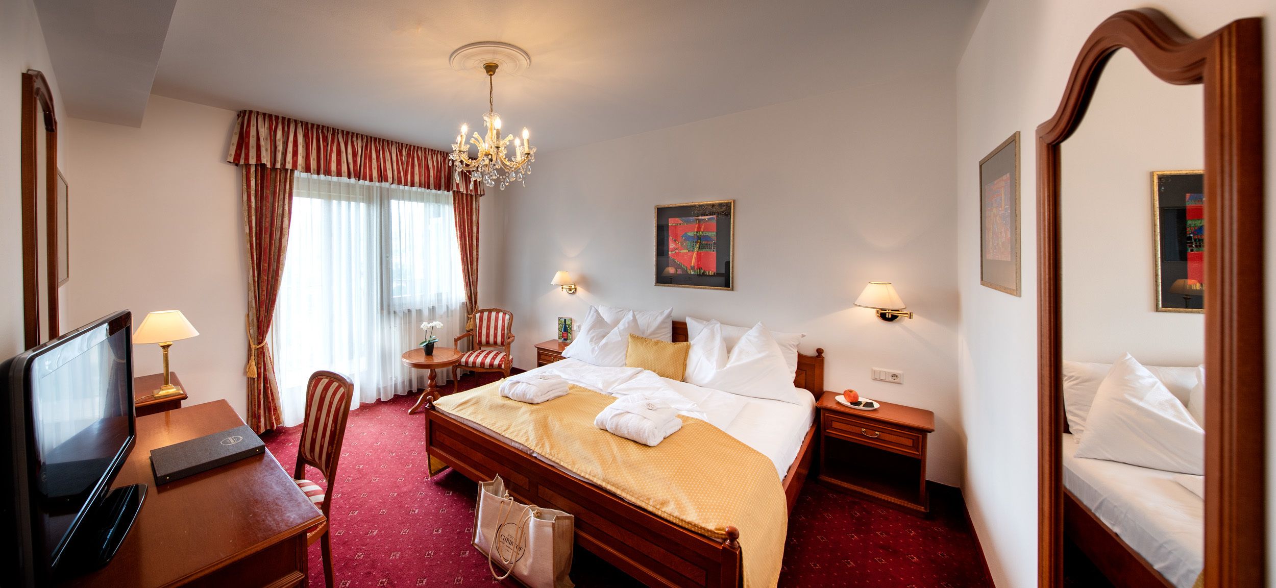 Ein Hotelzimmer mit einem Bett, einem Schreibtisch mit Fernseher, einem Sessel, einem kleinen Tisch mit Lampe und einer Kandelaber.