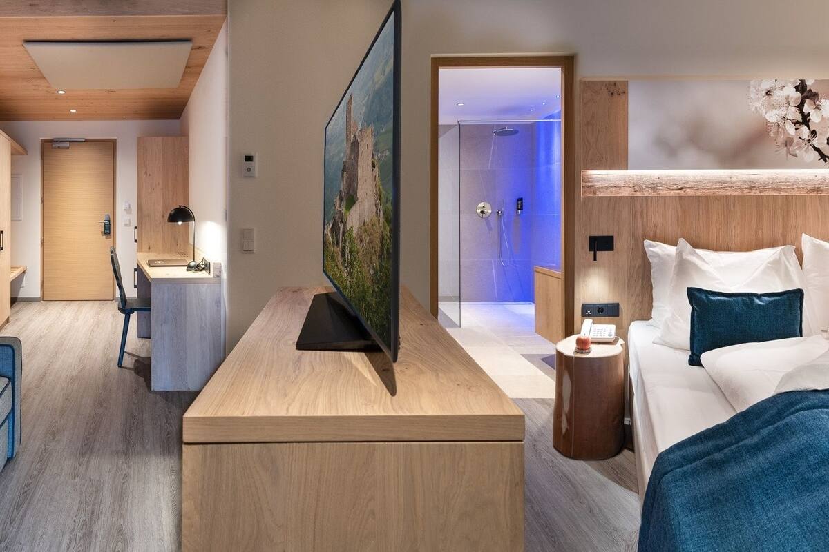 Une chambre d’hôtel moderne avec un plafond en bois, un grand lit avec une literie blanche et bleue, un bureau avec un ordinateur, un canapé avec des coussins bleus et un espace douche.