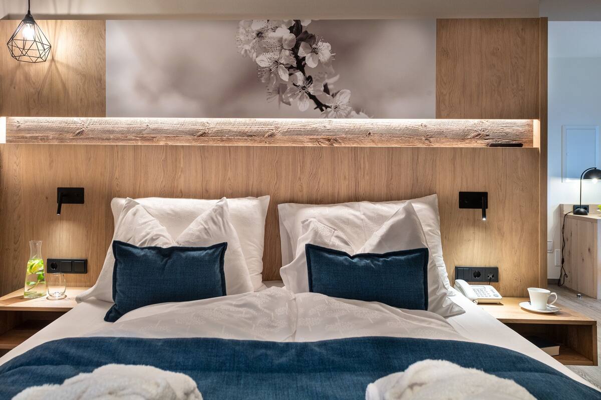 Une chambre d’hôtel moderne avec une tête de lit en bois, deux oreillers blancs, un plaid bleu, une table de chevet avec une lampe et un téléphone fixé au mur.