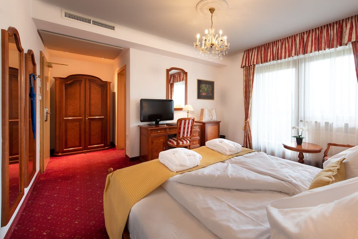 Une chambre d’hôtel avec un grand lit, un bureau avec une télévision, une chaise, une petite table avec une plante et une fenêtre avec des rideaux.