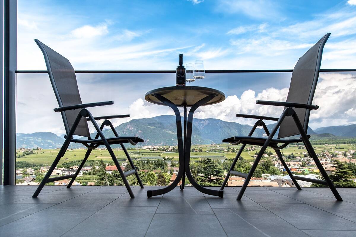 Une pièce dotée d’une grande fenêtre donnant sur une ville et des montagnes. On y trouve une table avec deux chaises et une bouteille de vin.
