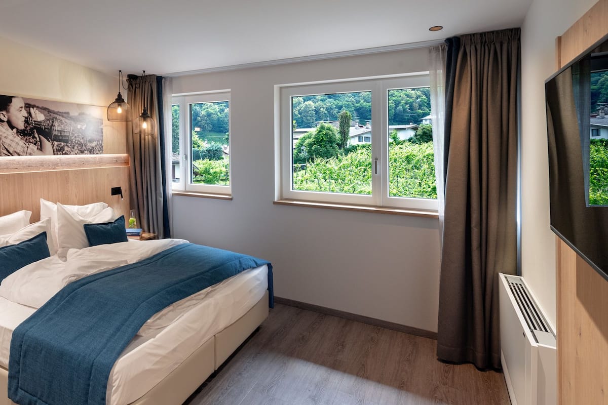 Une chambre d’hôtel moderne avec un grand lit, une tête de lit en bois, deux fenêtres avec des rideaux et une vue sur la verdure environnante.