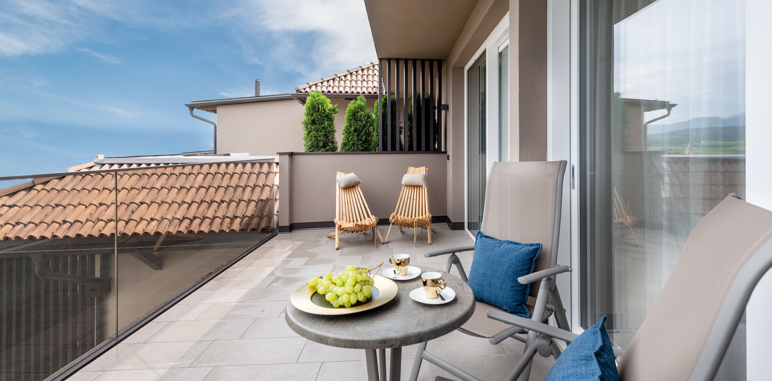 A balcony with a table set for two, featuring a plate of grapes and two cups. There are two wicker chairs and a cushioned chair. The balcony overlooks a building with a tiled roof and greenery.