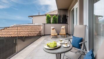 A balcony with a table set for two, featuring a plate of grapes and two cups. There are two wicker chairs and a cushioned chair. The balcony overlooks a building with a tiled roof and greenery.
