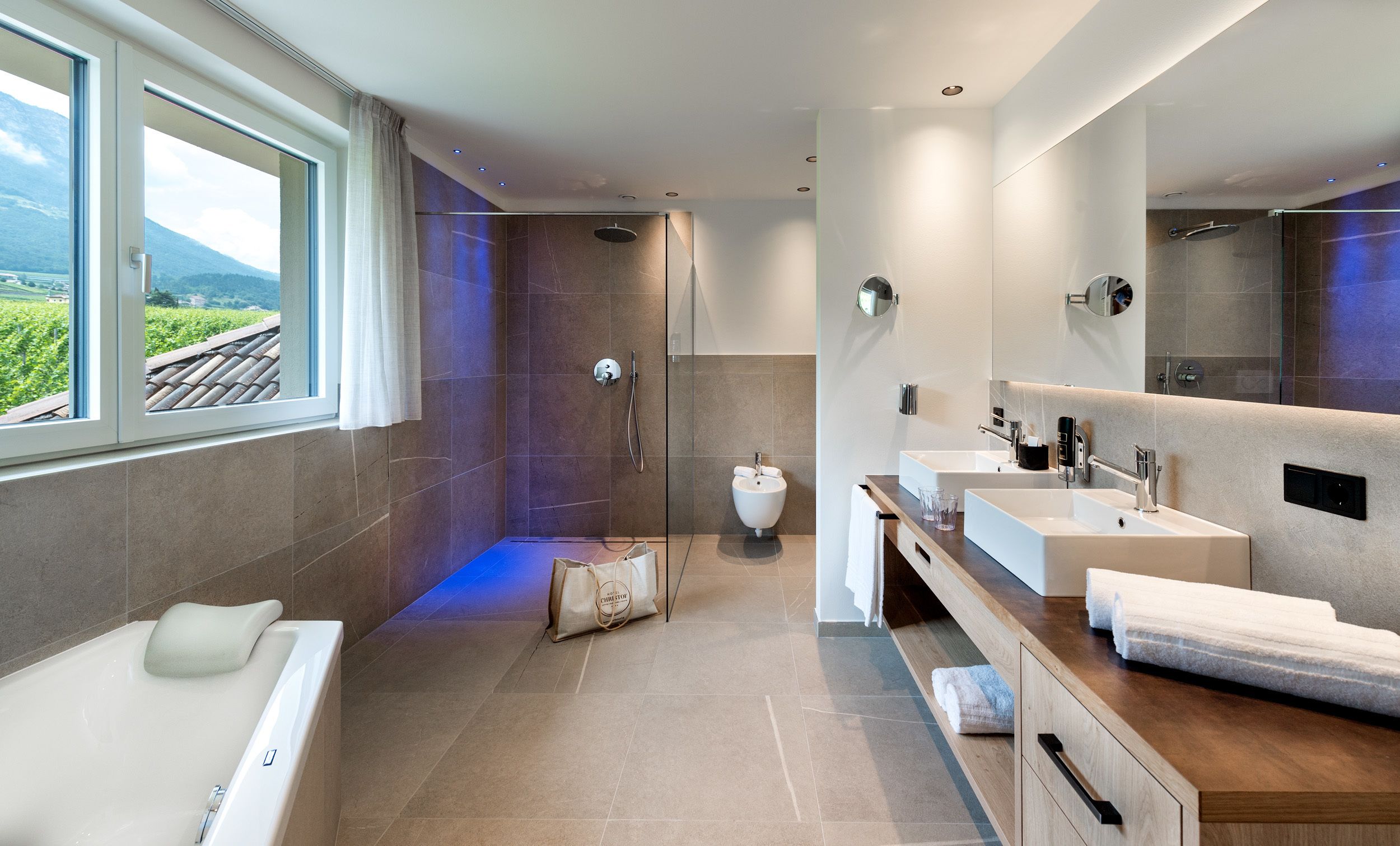Un baño moderno con bañera, dos lavamanos, ducha y un amplio ventanal con vistas a un paisaje montañoso.