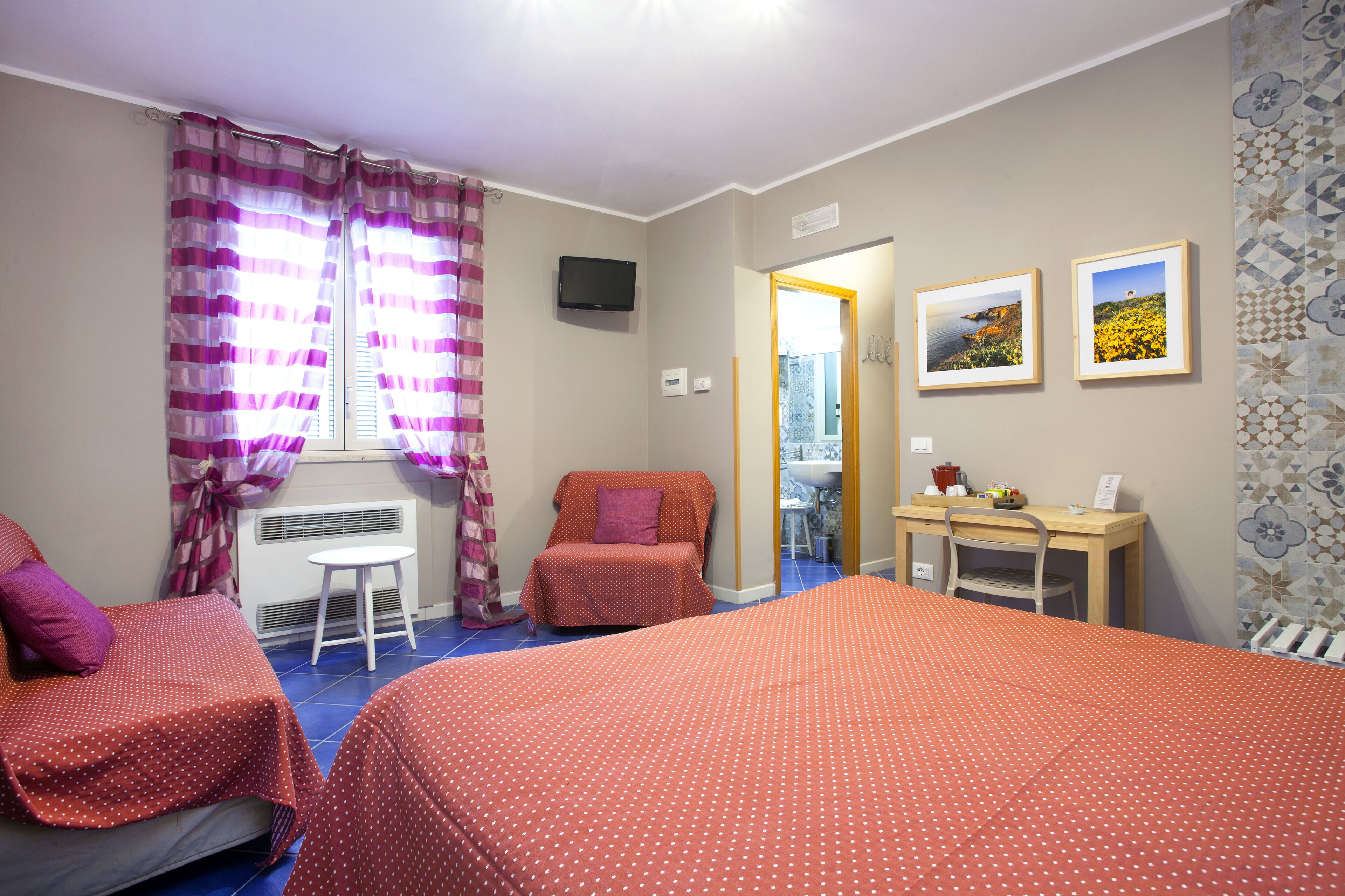 A bedroom with two beds, a desk, a chair, a radiator, a television, and a bathroom visible through an open door.