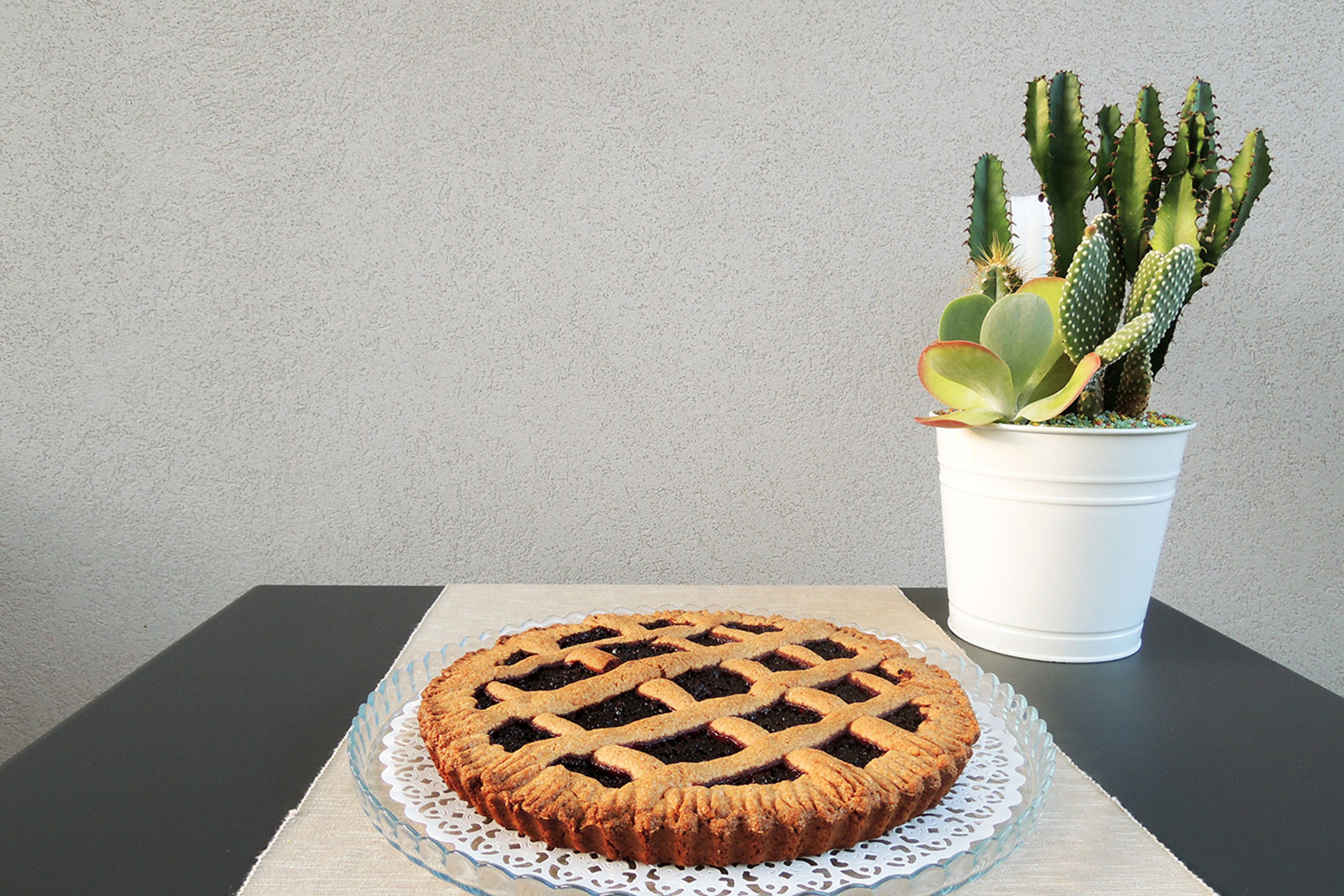 Une tarte ronde, posée sur une assiette en verre et recouverte d’une pâte en croisillons, est placée sur une table sur laquelle repose une nappe blanche en dentelle. À côté de la tarte, on aperçoit un pot blanc contenant une plante succulente, un cactus.