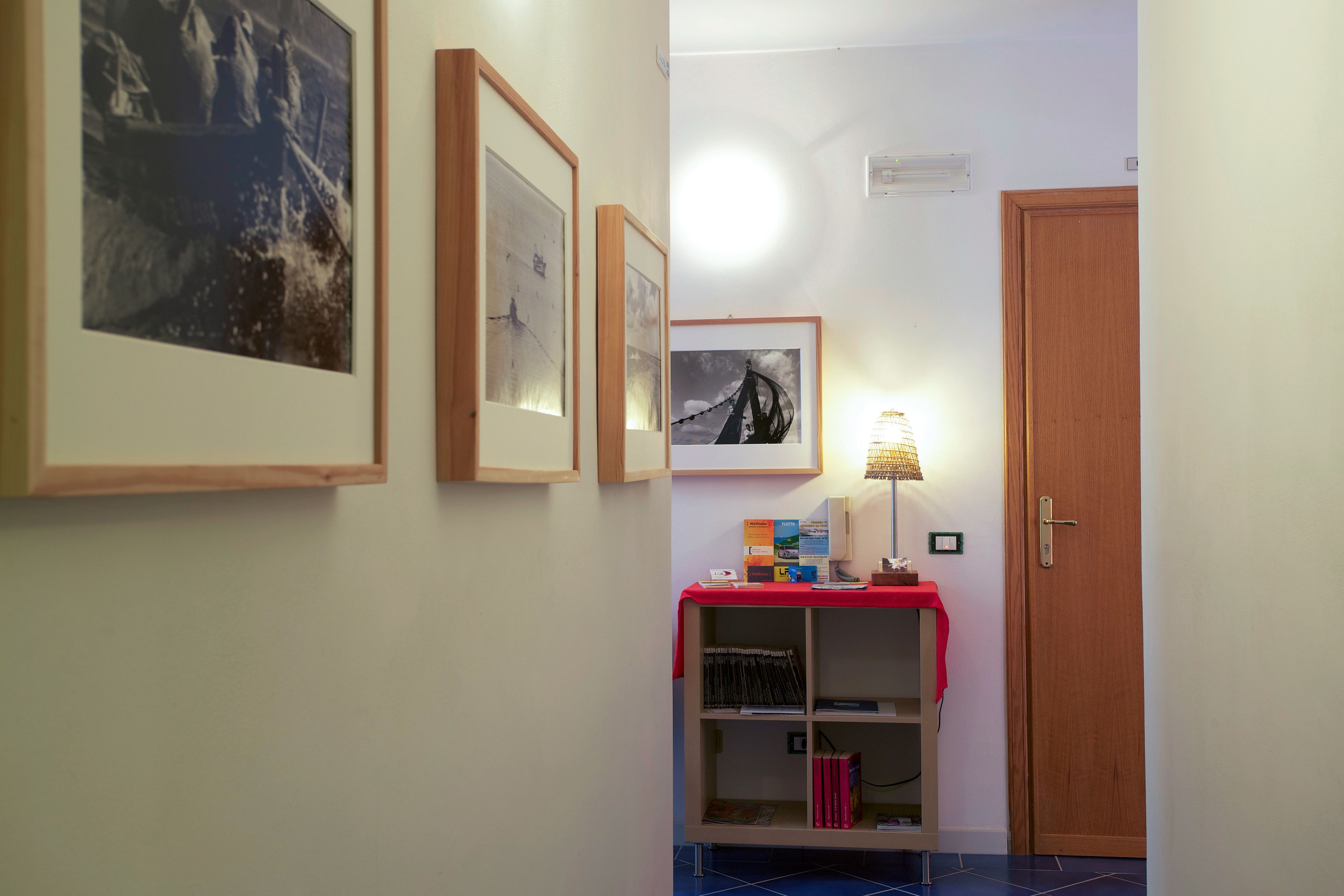 Een gang met ingelijste foto's aan de muur, een tafeltje met een lamp en een deur.