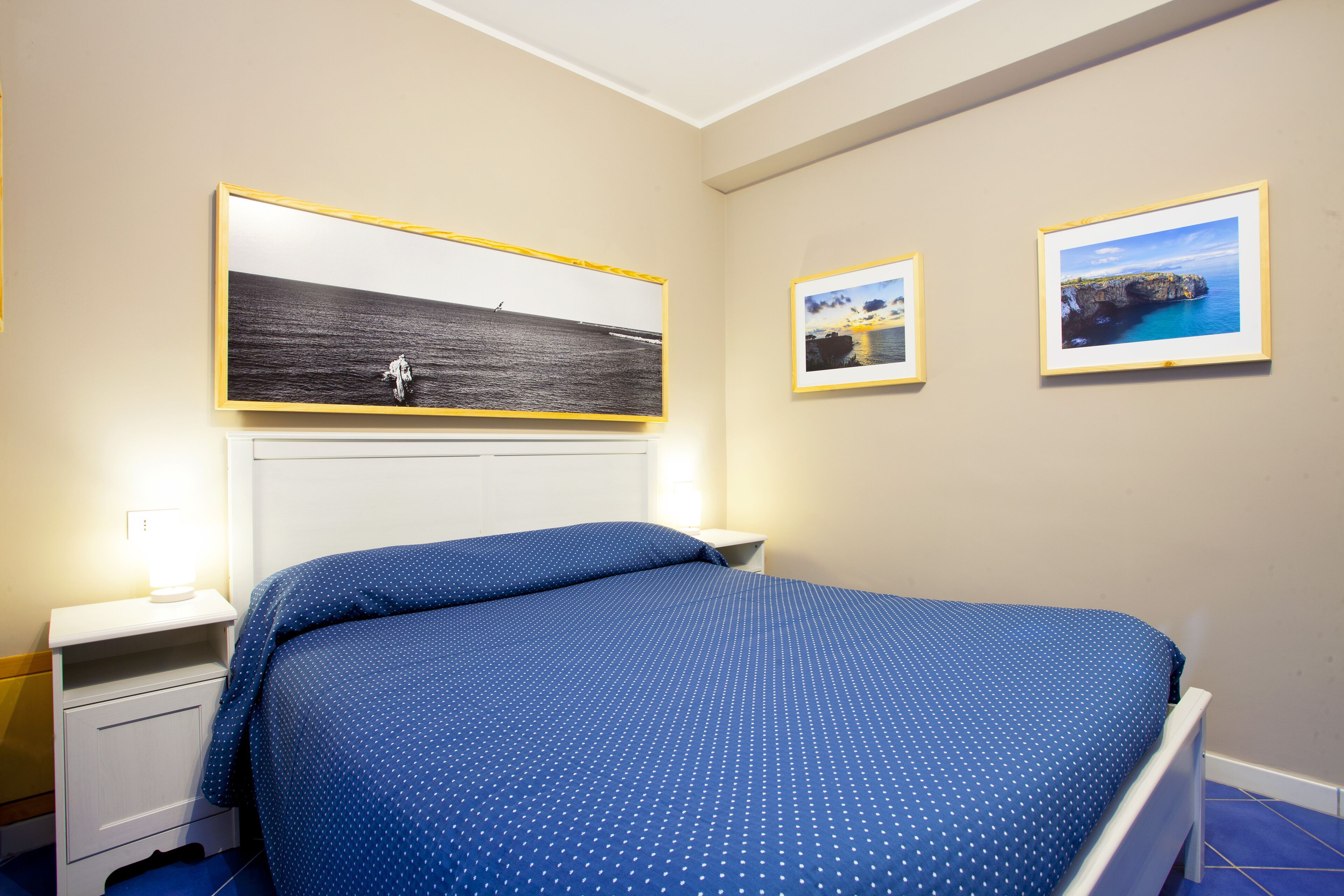 A bedroom with a bed, a nightstand, a wall-mounted shelf, and framed pictures on the wall.