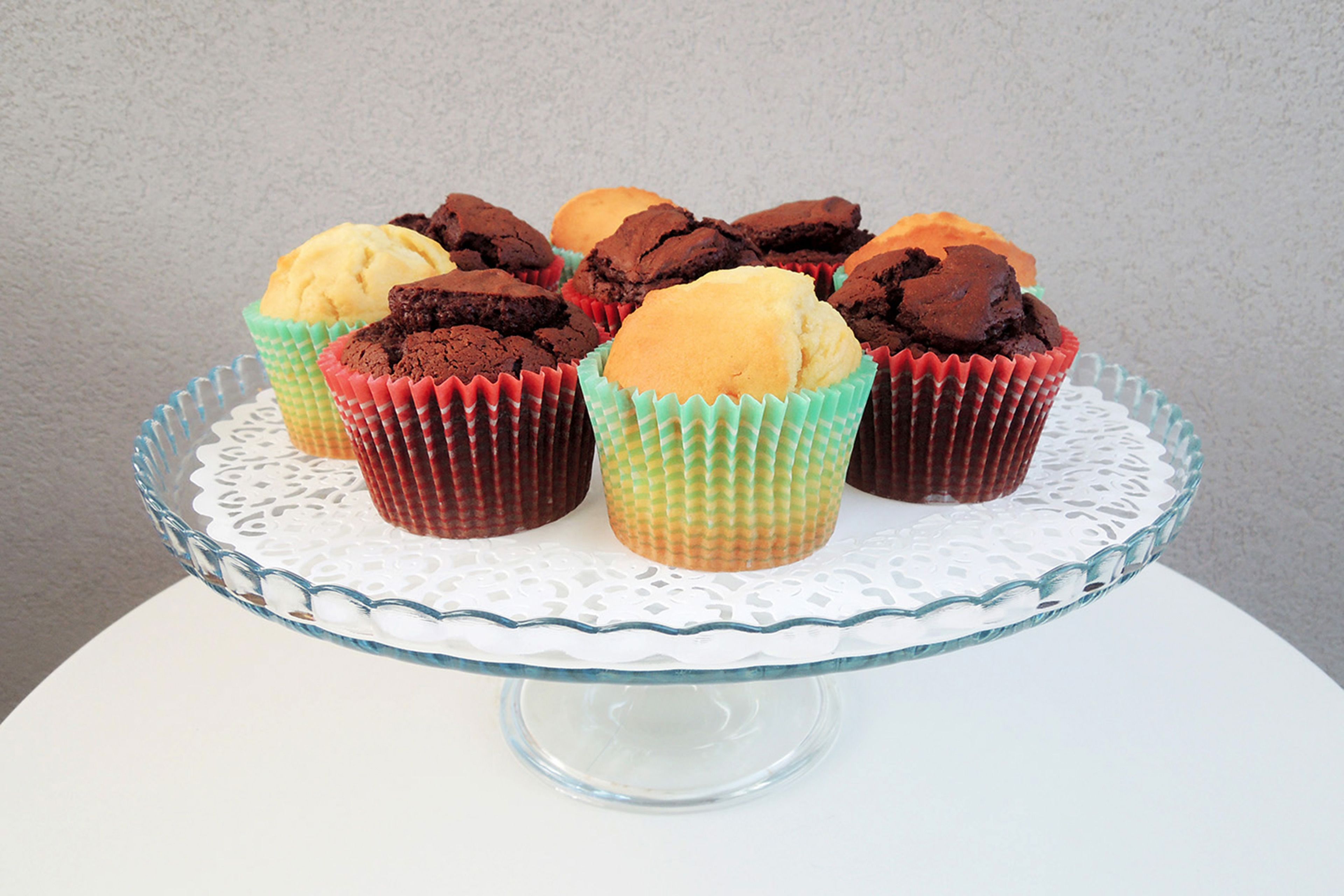 A clear glass cake stand with a lace doily base, holding several cupcakes in colorful paper liners.