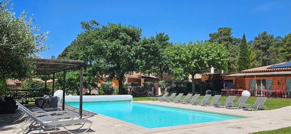 Piscina com espreguiçadeiras, pérgola e um edifício com telhado em azulejos.