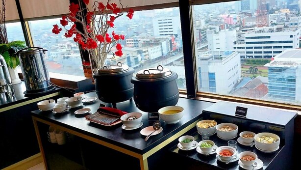 A kitchen counter with various dishes and cooking utensils, a window with a city view, and a vase with red flowers. - Grand Paragon (Jakarta)