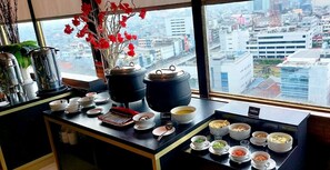 A kitchen counter with various dishes and cooking utensils, a window with a city view, and a vase with red flowers. - Grand Paragon (Jakarta)