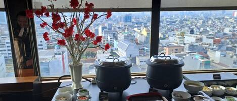 Una mesa de comedor con diversos platos y un jarrón de flores rojas, con vistas a un paisaje urbano.