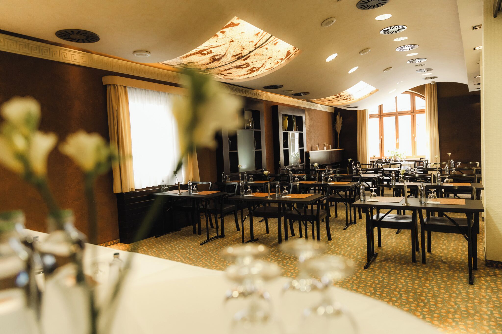 A hotel dining area with multiple tables and chairs, a large window with curtains, and a ceiling with recessed lighting.