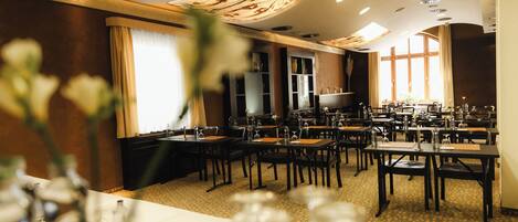 A hotel dining area with multiple tables and chairs, a large window with curtains, and a ceiling with recessed lighting.