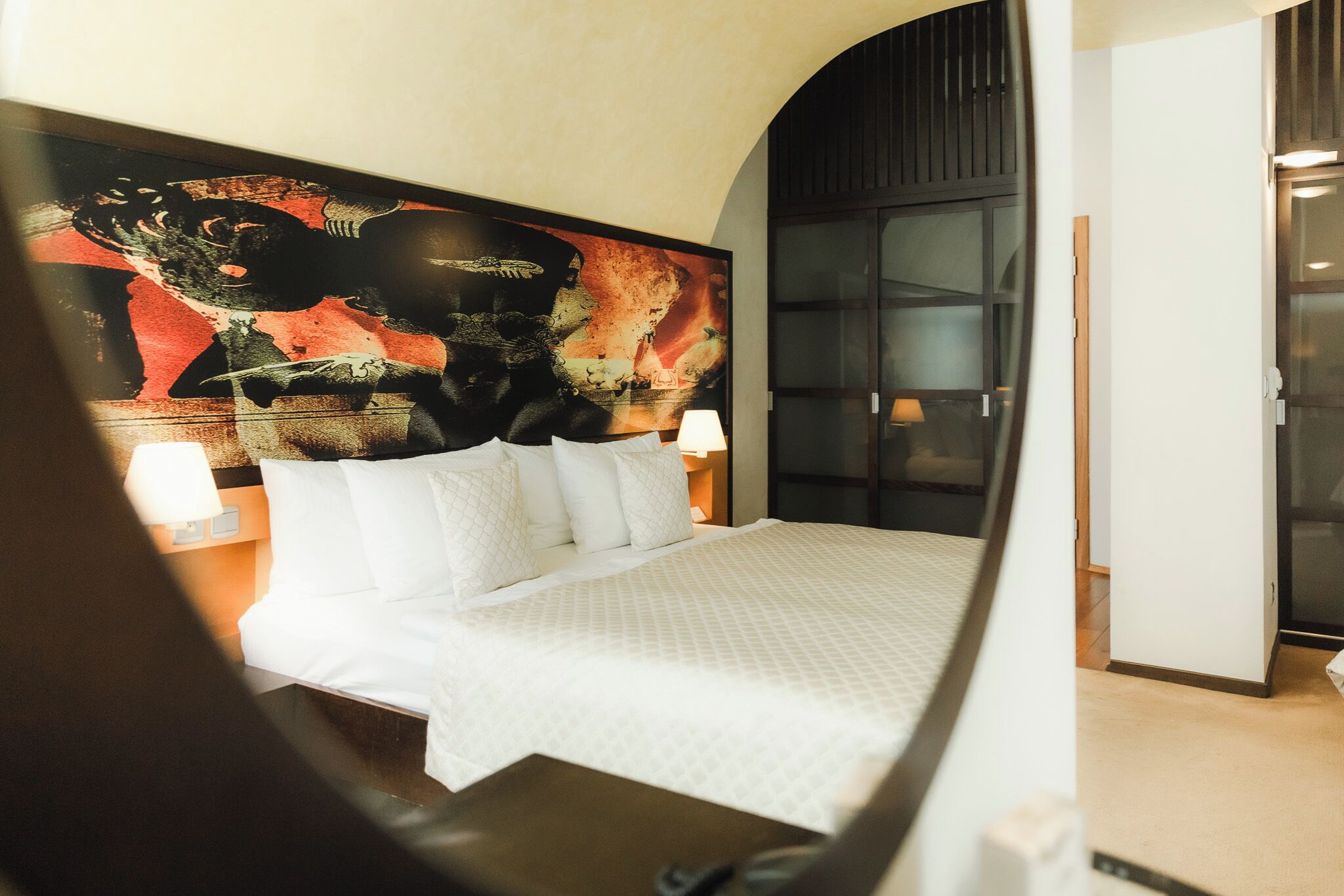 A hotel room with a large bed, a nightstand with a lamp, and a large abstract painting on the wall.