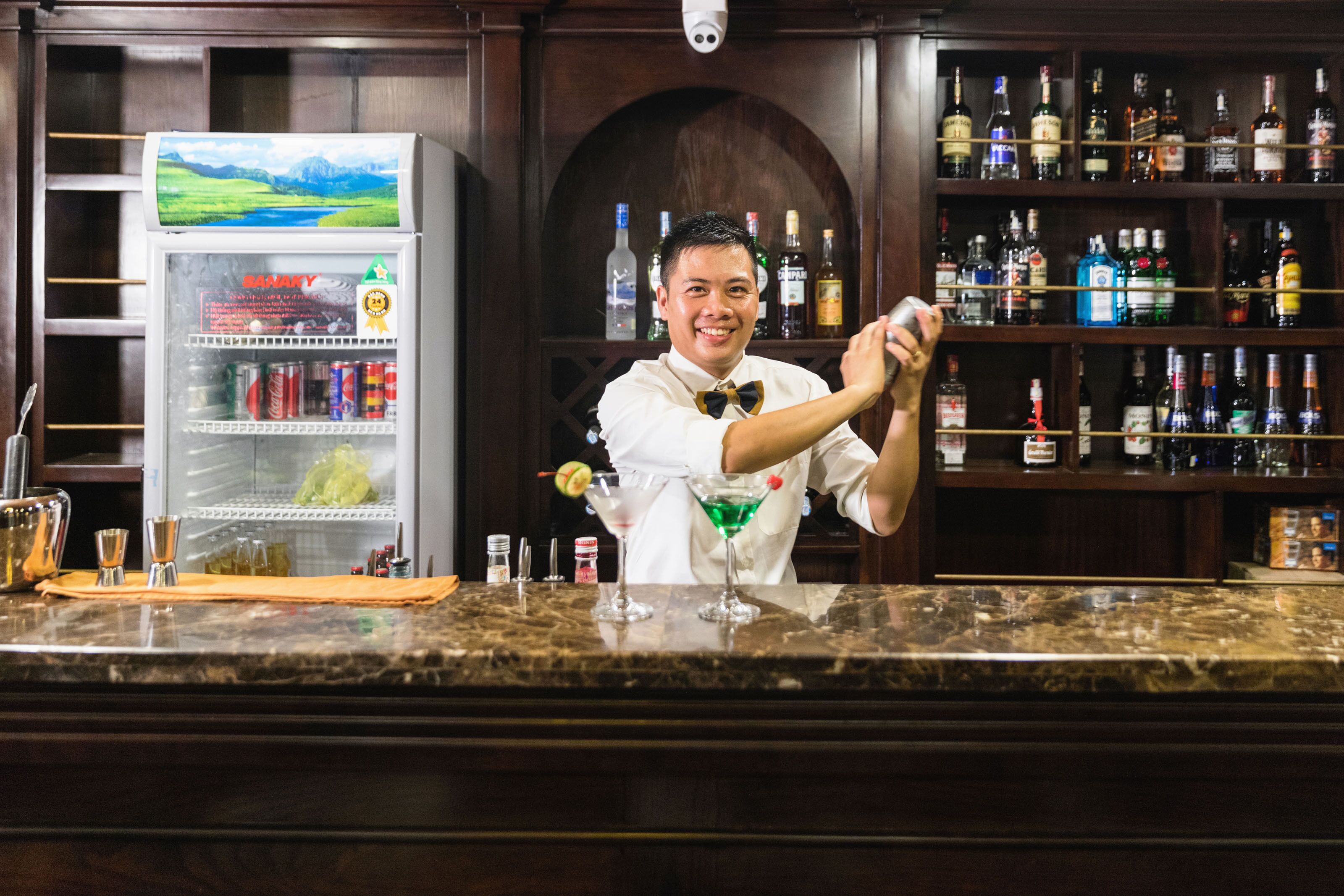 A bar area with a bartender preparing a drink, a minibar, and shelves of liquor bottles.