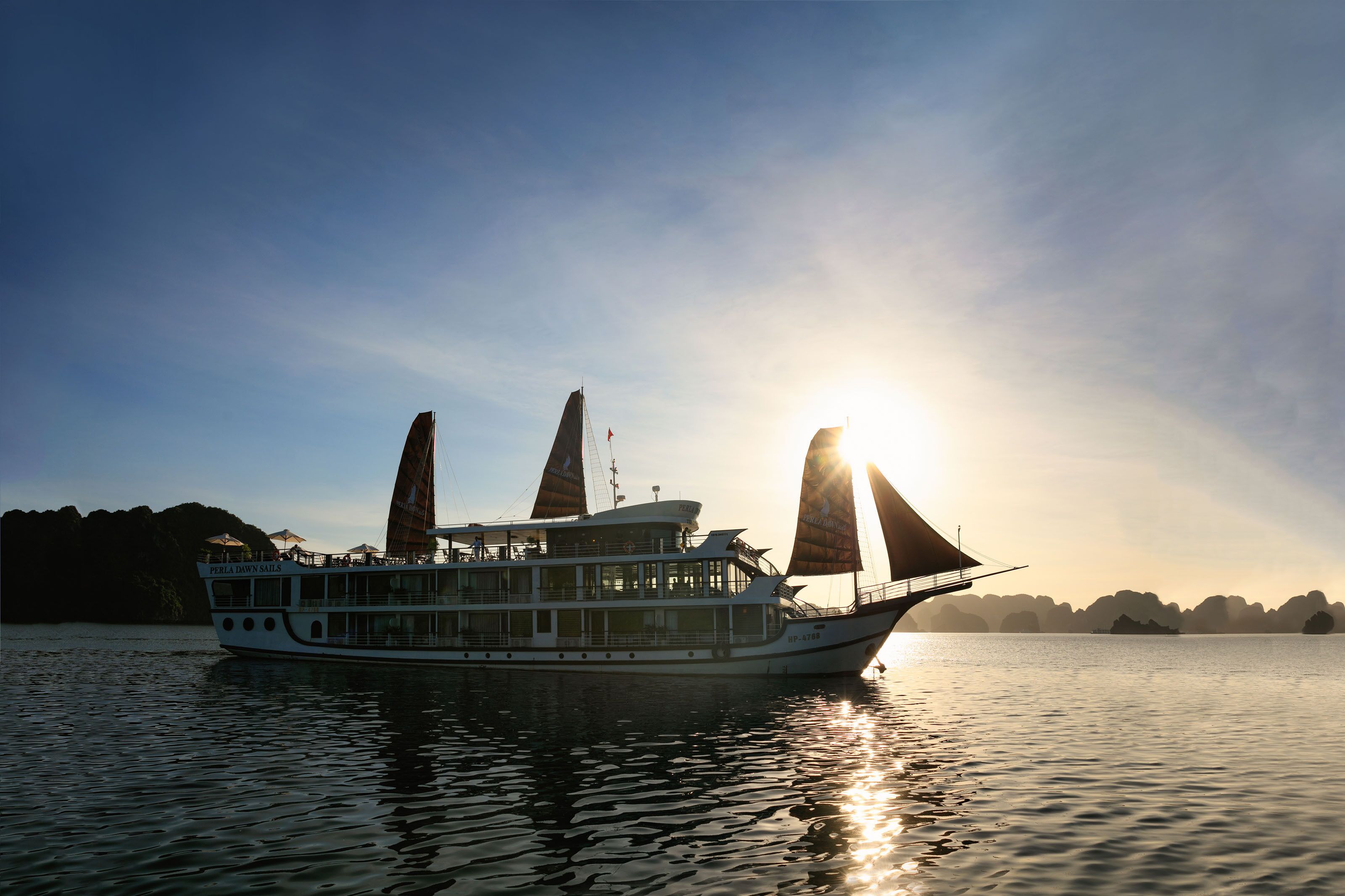 Ein Schiff mit mehreren Decks und einer markanten Segelkonstruktion, das auf ruhigem Wasser unter einem wolkenlosen Himmel mit entfernten Landformen fährt.
