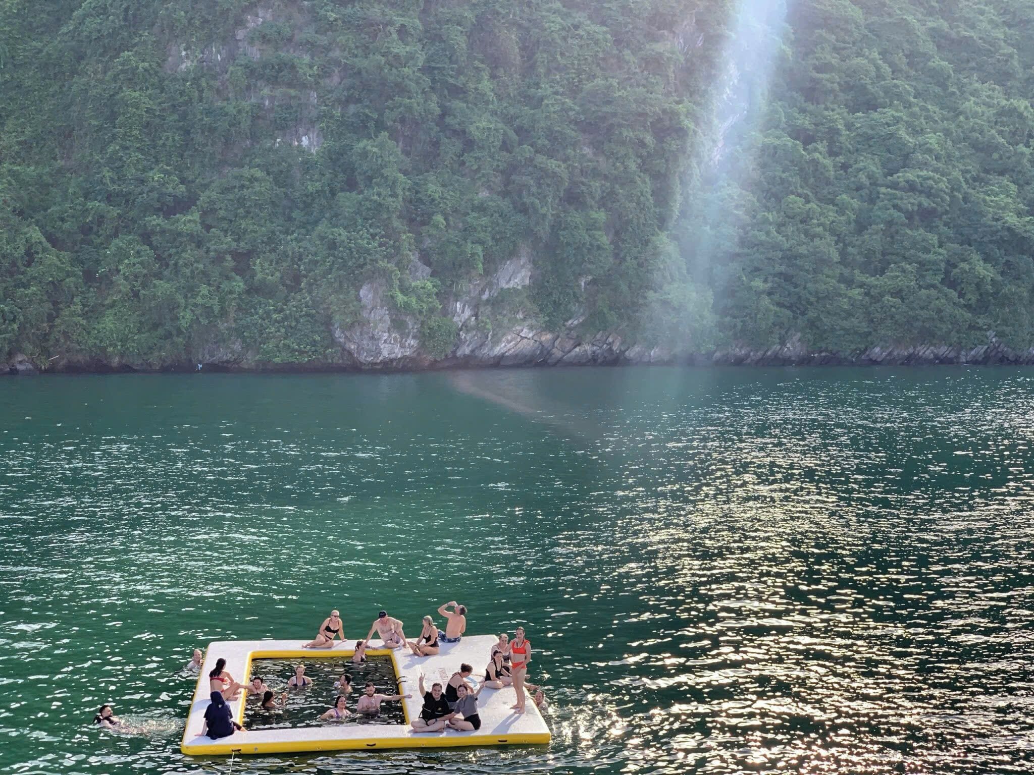 Un grupo de personas en una plataforma flotante sobre el agua, con un acantilado cubierto de bosque al fondo.