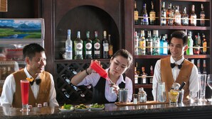 A bar area with bartenders preparing drinks, shelves stocked with liquor bottles, and a menu board in the background. - V'Spirit Cruises (Tuan Chau)