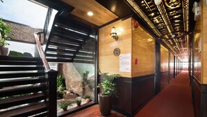 A hotel corridor with wooden flooring, a red carpet, a wooden wall with a light fixture, and a potted plant. - V'Spirit Cruises (Tuan Chau)