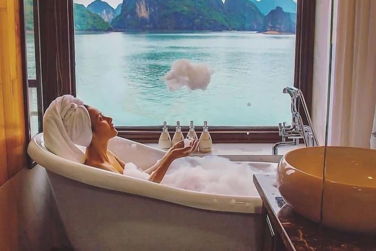 Una bañera con vistas a un cuerpo de agua tranquilo y montañas, una toalla en la cabeza, y botellas de champú y acondicionador.