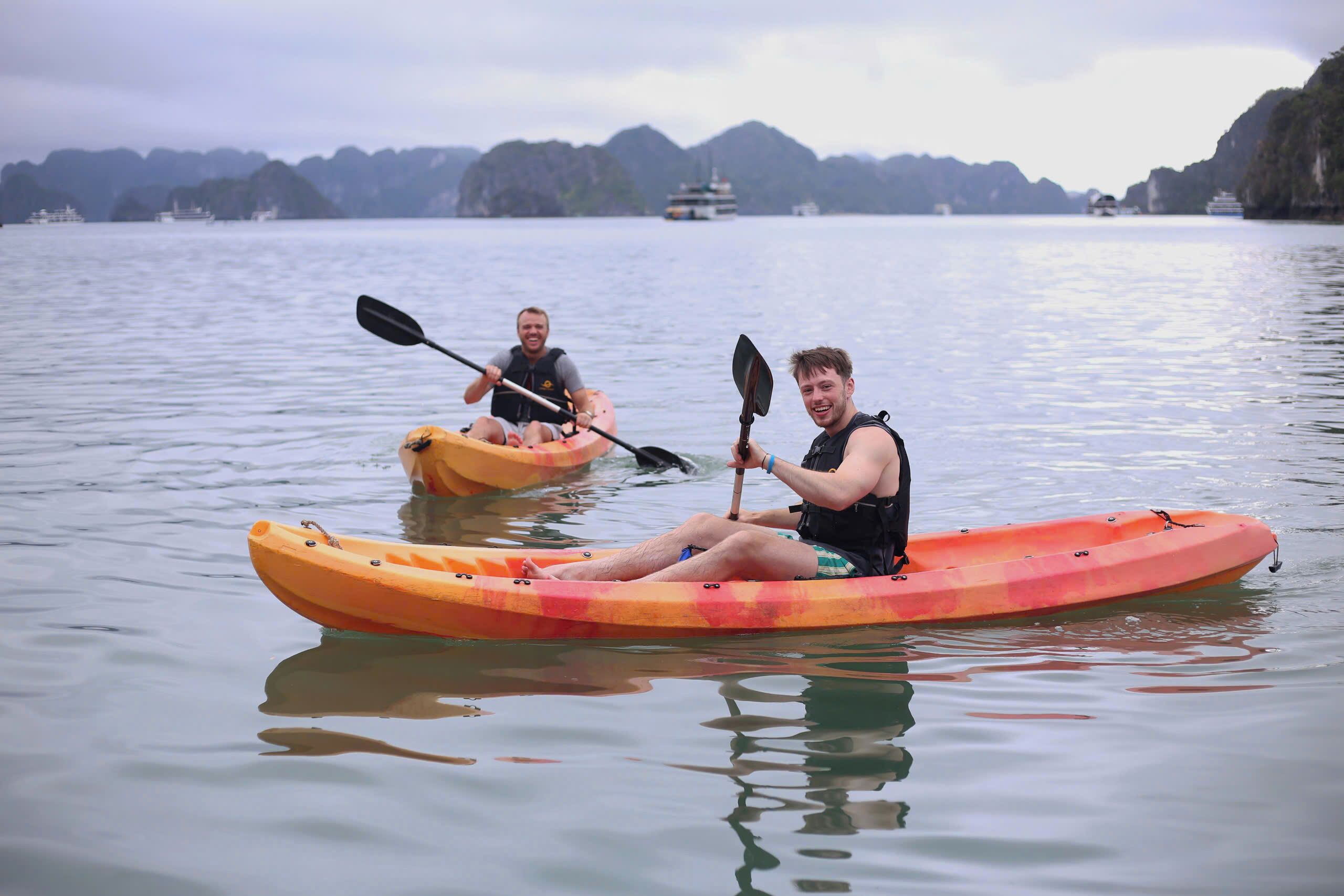 Deux personnes pratiquent le kayak sur une eau calme, avec des bateaux en arrière-plan.